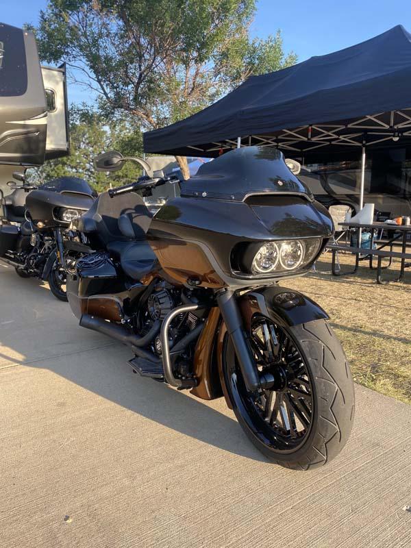 A parked motorcycle under a black pop-up canopy, with picnic tables and trees in the background.